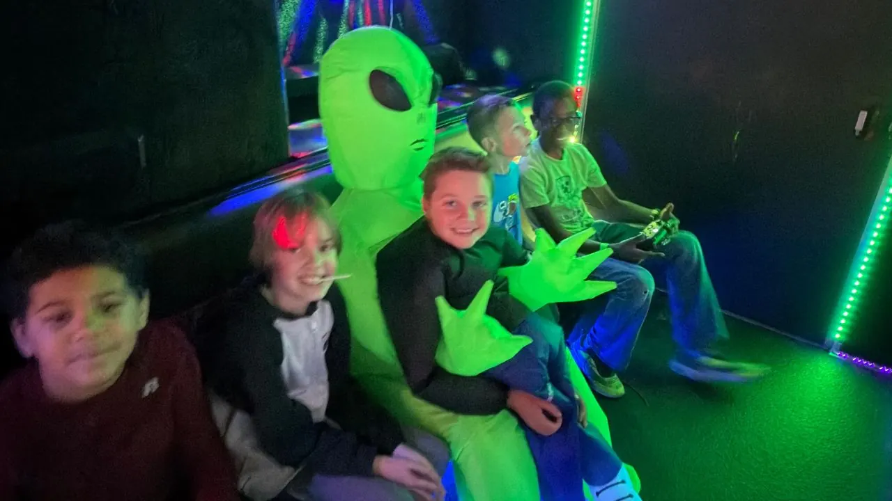 Kids posing with a green alien mascot inside the neon-lit mobile gaming truck during a themed birthday party