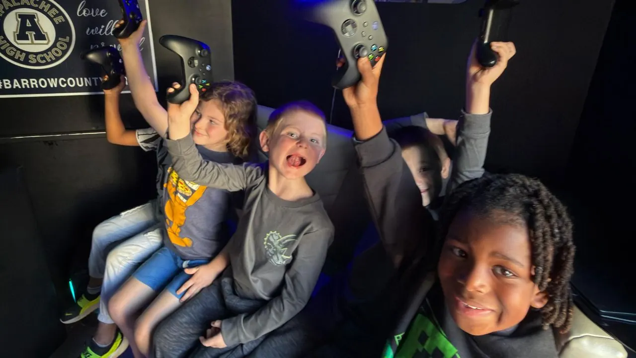 Four kids holding Xbox and PlayStation controllers in the air celebrating inside the Ultimate Mobile Gaming truck at a school event